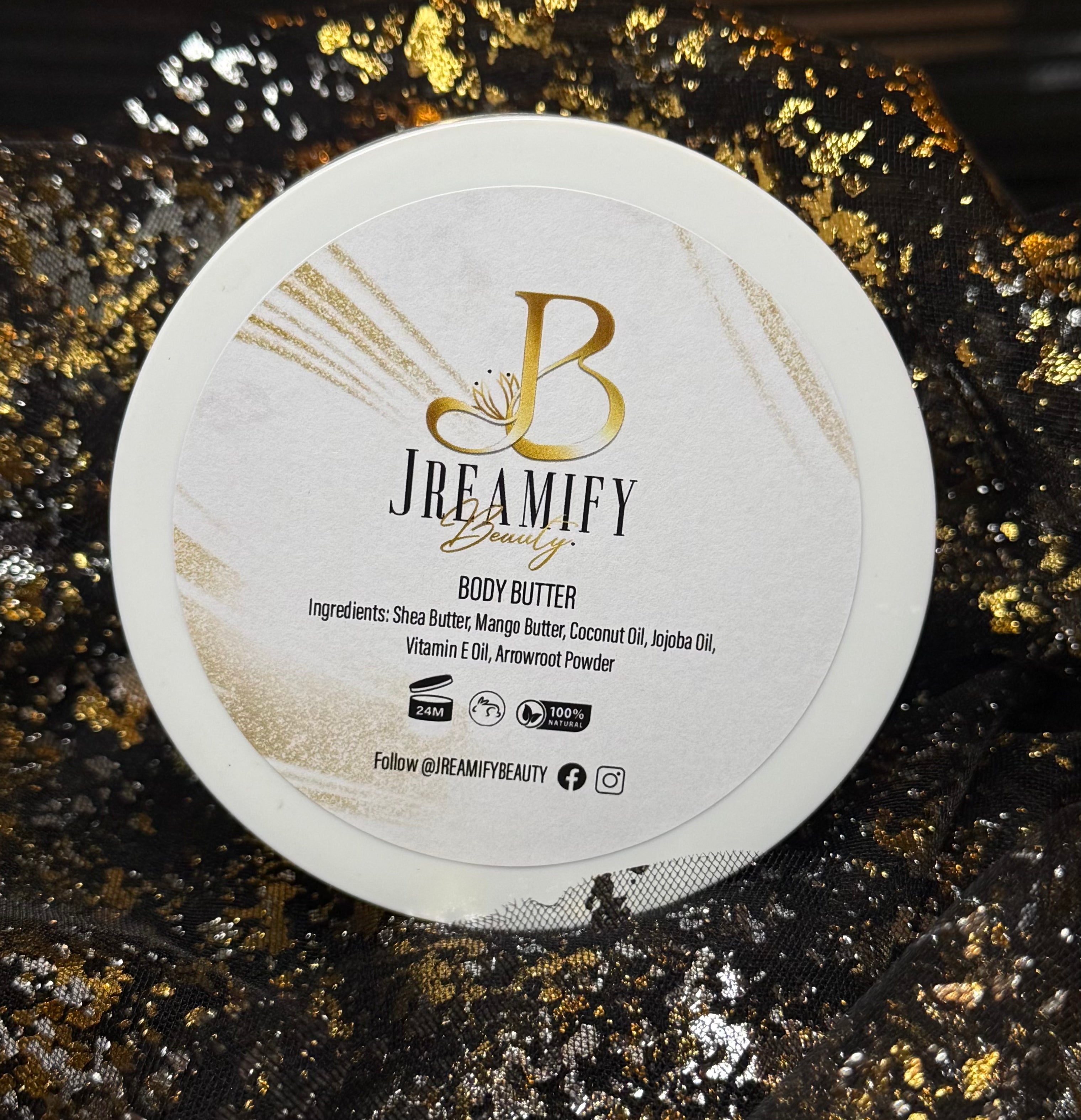 A round white tub of Jreamify Beauty Body Butter with a white lid featuring gold and black text, set against a sparkling black and gold sequined fabric background.