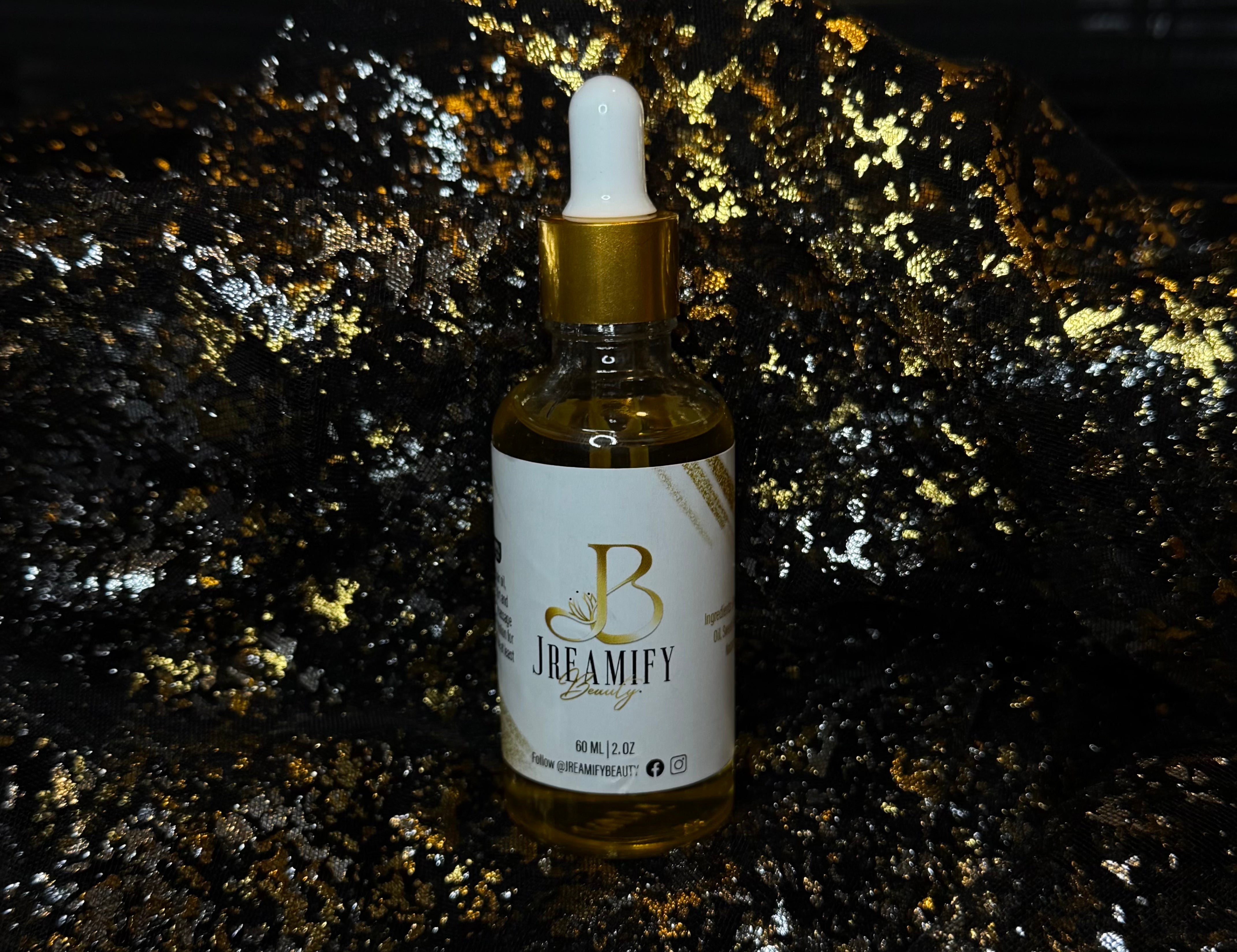 A bottle of Jreamify Beauty hair oil with a white dropper cap and gold collar, set against a dark background with gold glitter.