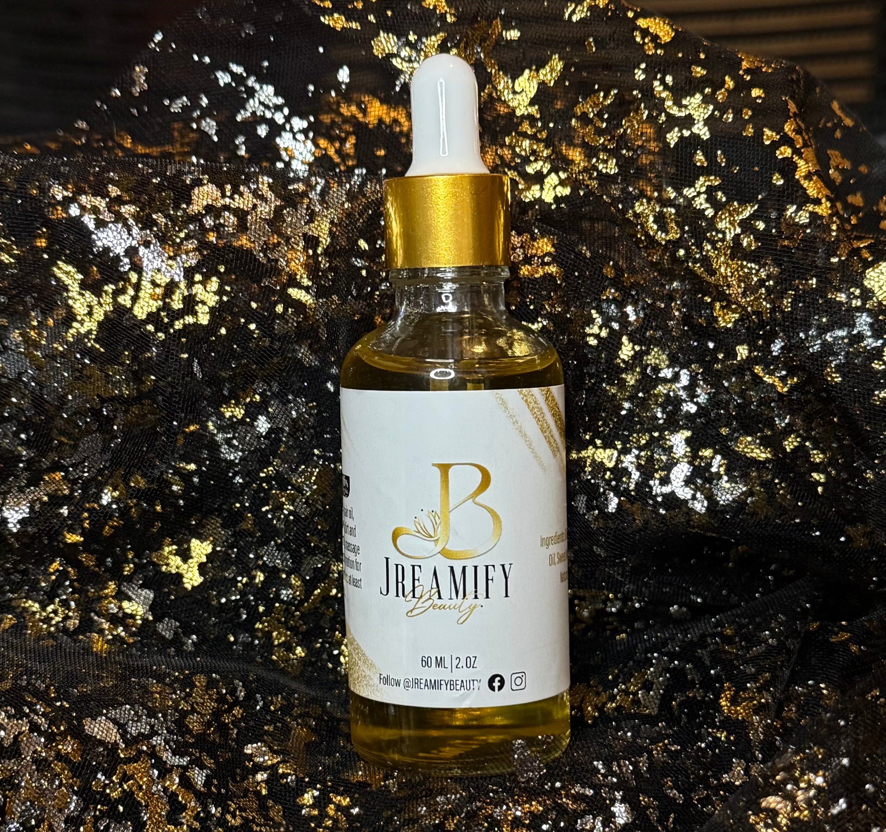 A bottle of Jreamify Beauty hair oil with a white dropper cap and gold collar, set against a black sequined fabric background with gold glitter.