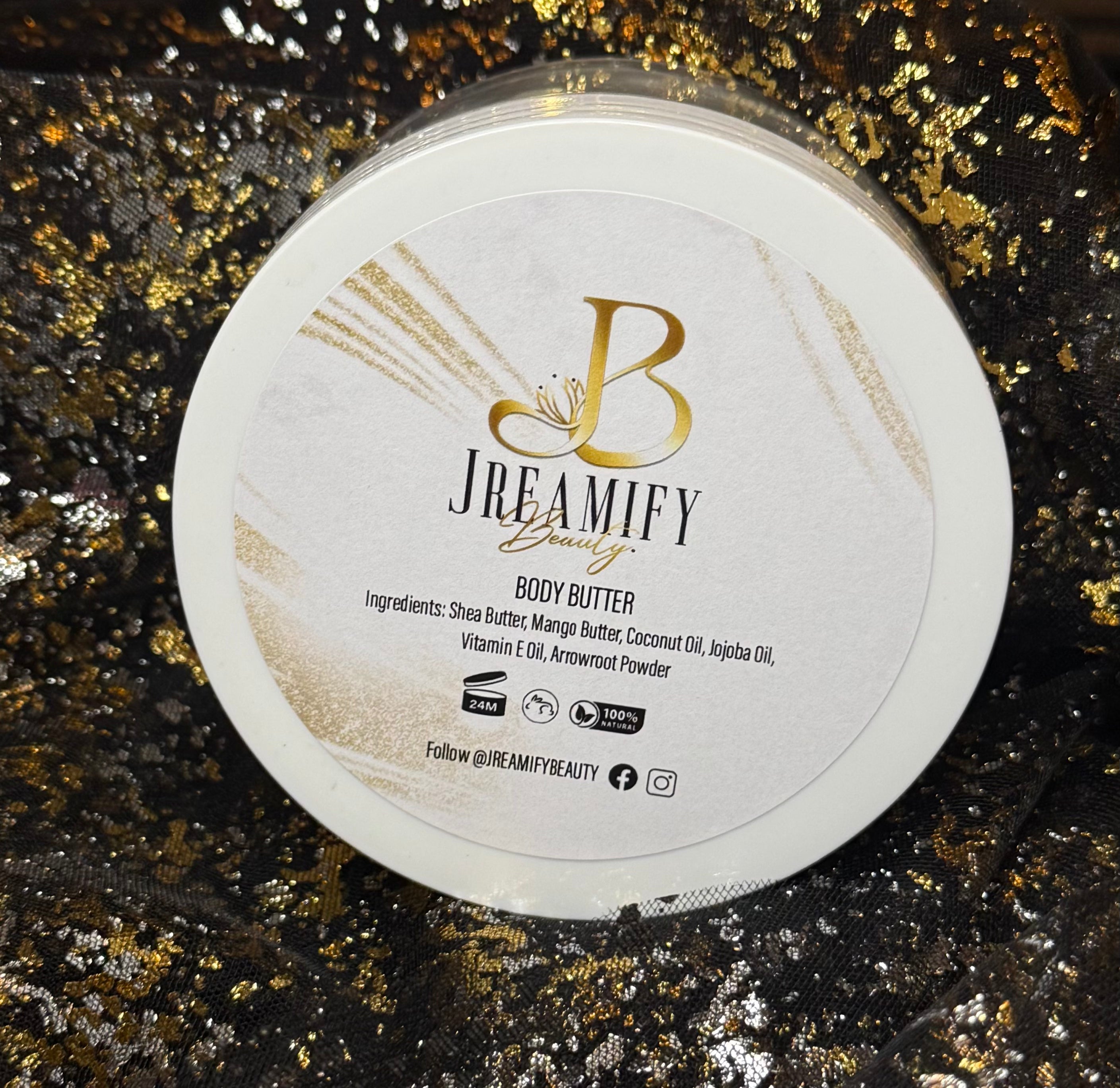 A round white jar lid for Jreamify Beauty Body Butter, featuring a white and gold label with a prominent gold 'B' logo, set against a black and gold glittery background.