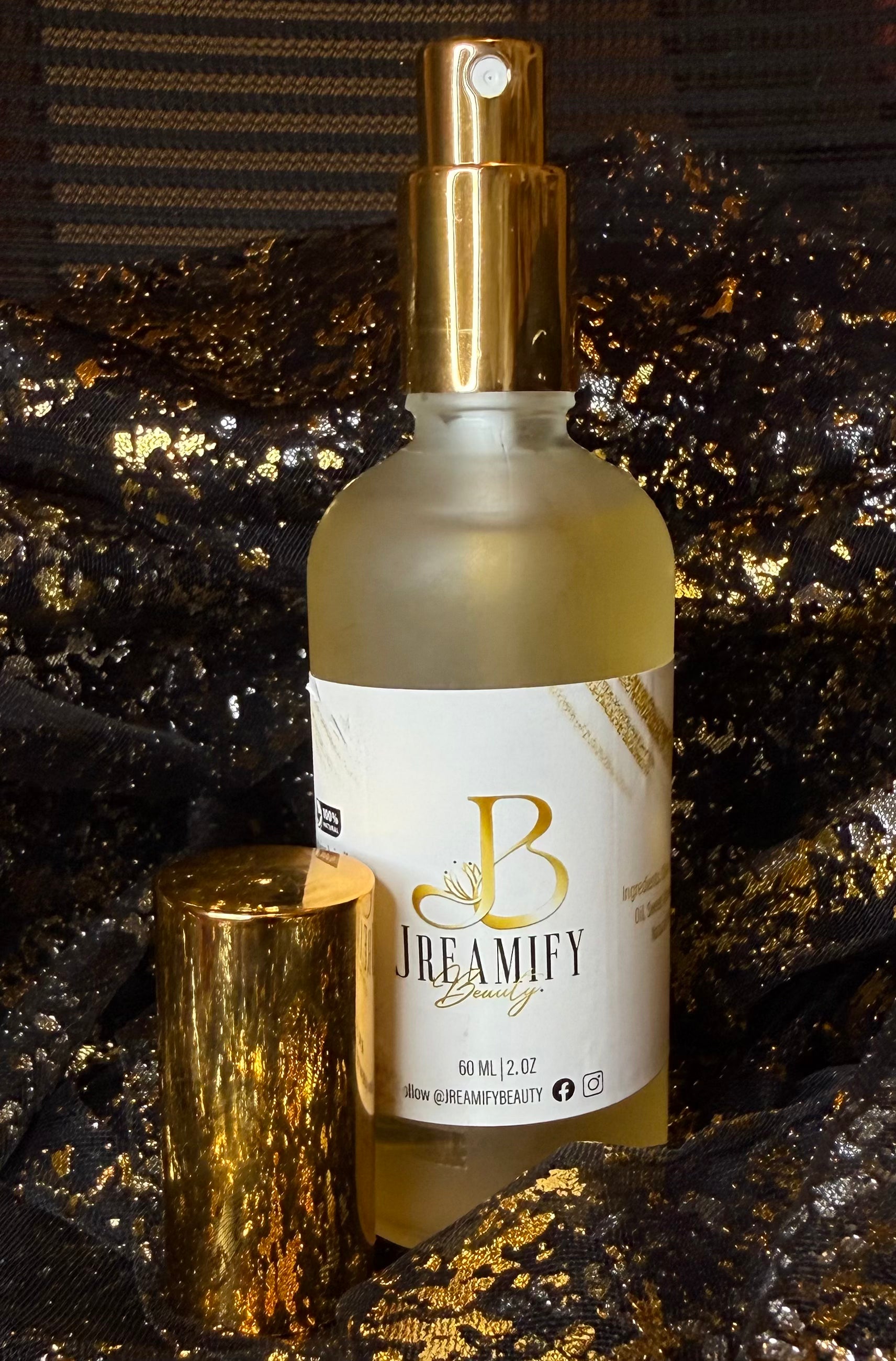 Jreamify Beauty hair product bottle with a gold spray top and white label, shown with its gold cap beside it on a glittery black and gold textured background.
