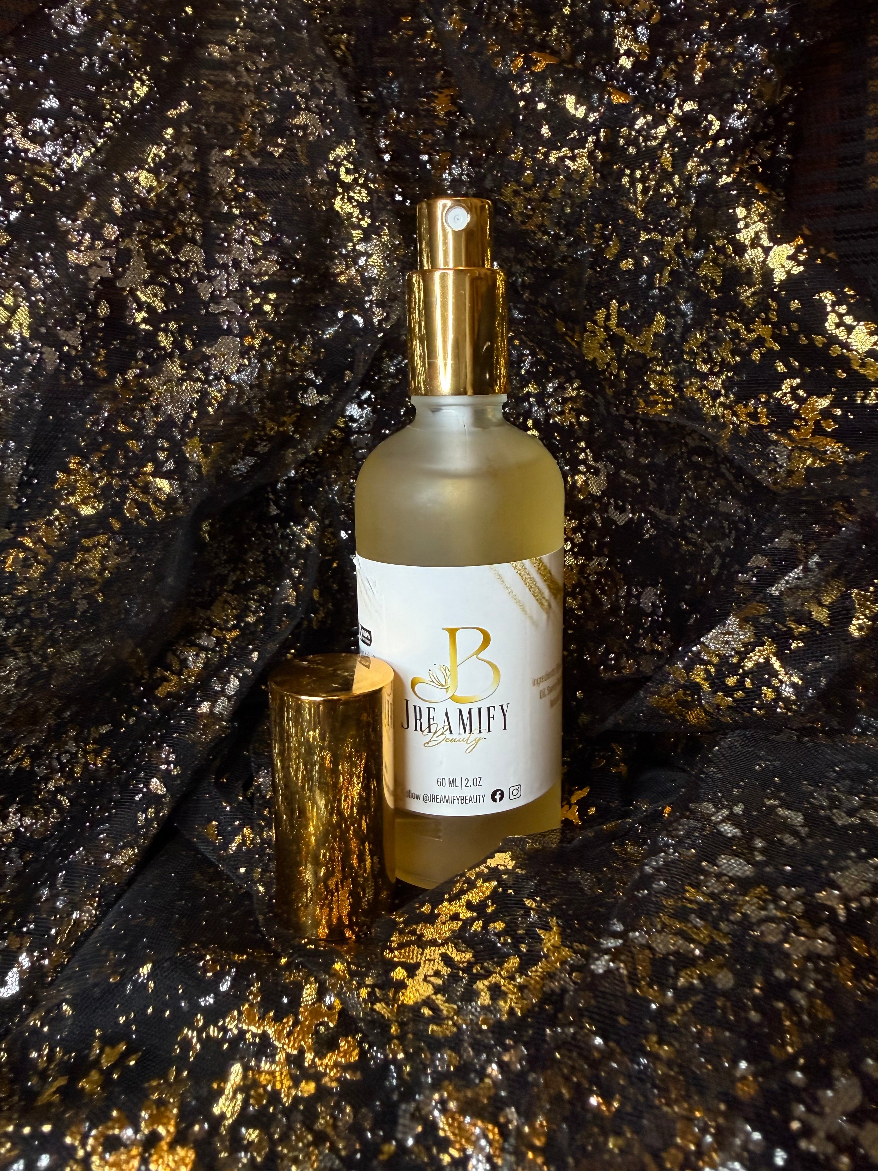 A frosted glass spray bottle with a gold pump dispenser and a white label reading 'Jreamify Beauty', shown with its gold cap beside it on a glittery black and gold surface.